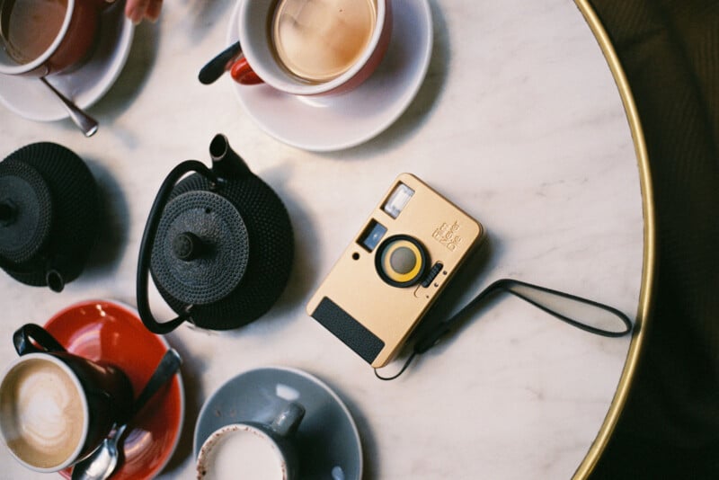 A top-down view of a marble table with two black teapots, a gold vintage camera, and three cups of coffee with varying amounts of milk, in red and gray saucers. A hand holds a cup at the top left corner.