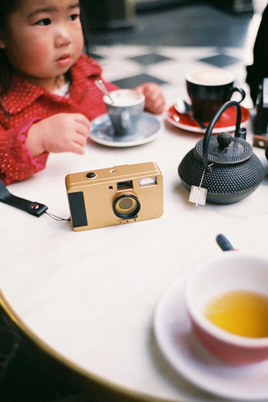 A child in a red outfit sits at a white table with a teacup, black teapot, and yellow camera. The background shows a black and white checkered floor.