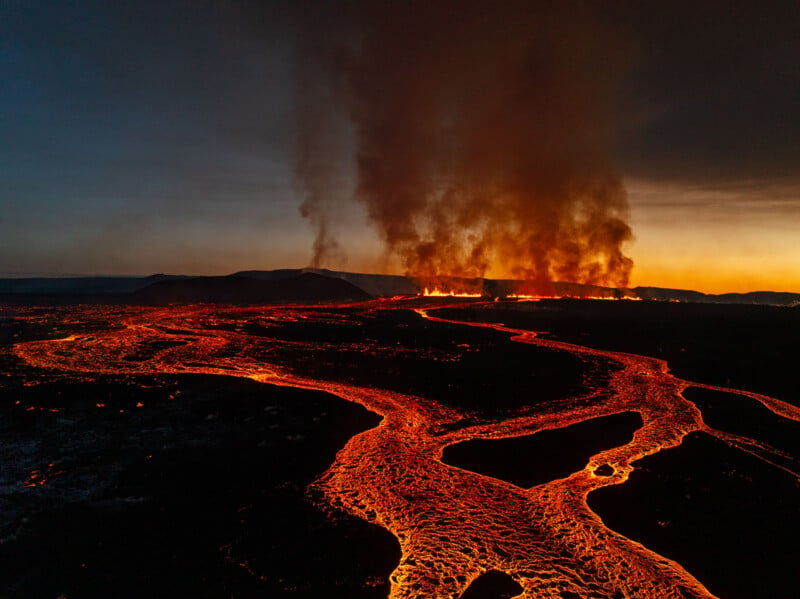 Aerial view of a volcanic eruption at night, with glowing rivers of lava flowing across dark terrain. Thick plumes of smoke rise into the sky, illuminated by the fiery glow of the molten lava.