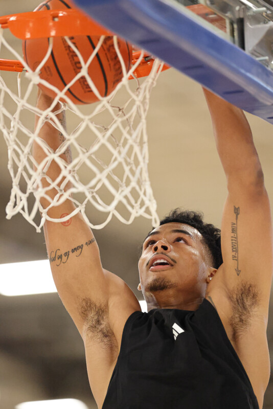 A basketball player in a black jersey jumps to perform a slam dunk. The ball is just about to pass through the hoop. Tattoos are visible on his arms, and he focuses intently on the basket.