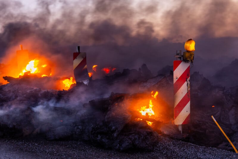 A scene of a dark landscape with glowing orange flames and thick smoke rising from burning rubble. Two striped warning signs with a yellow flashing light stand amidst the debris, indicating caution.