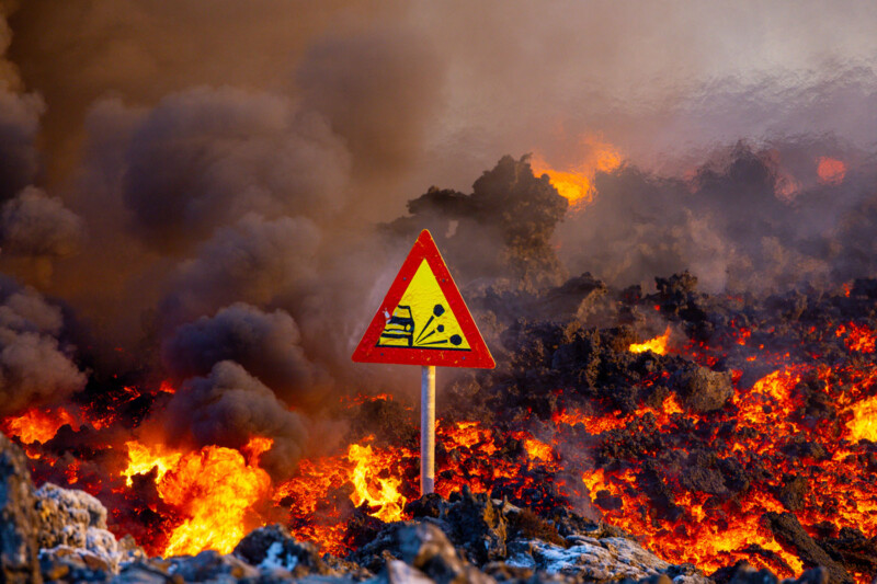 A road sign with the warning of falling debris stands amidst a landscape filled with smoke, fire, and molten lava, indicating a volcanic eruption or intense fire nearby.