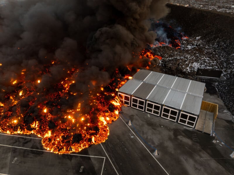 Aerial view of a large fire with thick black smoke billowing over an industrial area. Flames spread near a rectangular building. The parking lot and some empty spaces are visible around the structure.