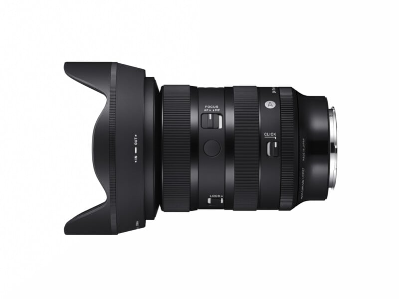 A black camera zoom lens with multiple dials and switches on its body, including focus and lock controls. The lens hood is attached. The lens has markings showing focal length and aperture settings. The brand logo is visible.