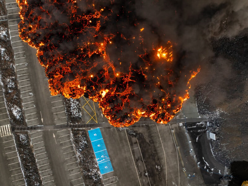 Aerial view of a large, intense fire with thick black smoke spreading across a parking lot. Visible blue and white handicapped parking spaces near the fire. Surrounding areas appear empty, with ash and debris scattered.