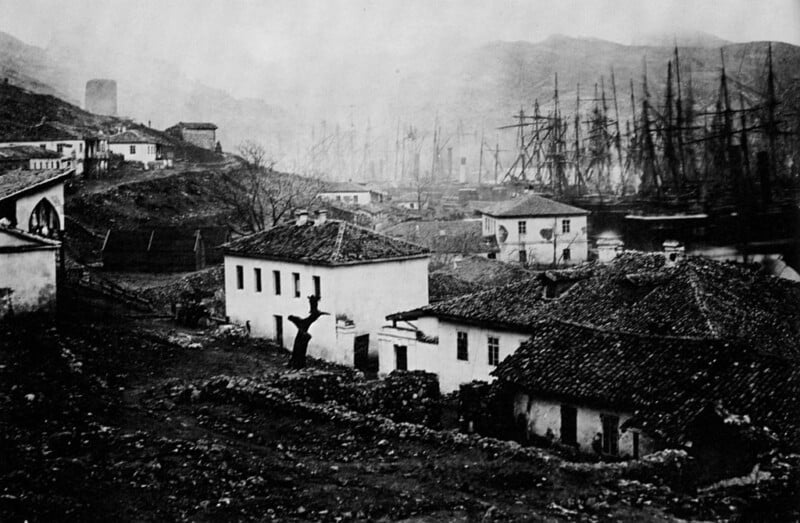 A black and white photograph depicting a historical harbor scene with several sailing ships docked. In the foreground, there are dark-roofed buildings and a tree. Hills are visible in the misty background.