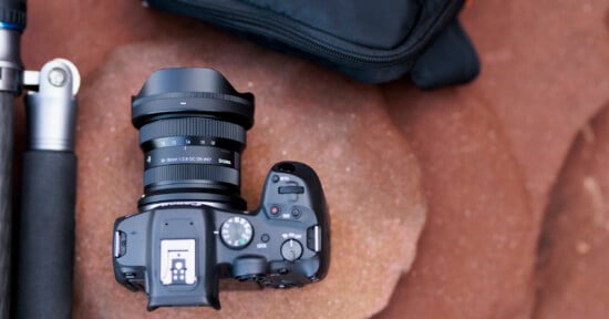 A black digital camera with a wide-angle lens sits on a reddish-brown rock surface. To the left is the leg of a tripod, and above the camera is a partially visible black camera bag. The camera controls and lens details are visible.