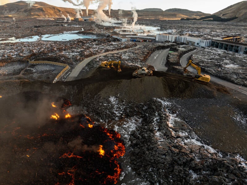 An aerial view of an erupting volcano with flowing lava near a road. Heavy machinery is seen managing the lava flow, while smoke rises in the background. The landscape is rugged and sparse, under a clear sky.