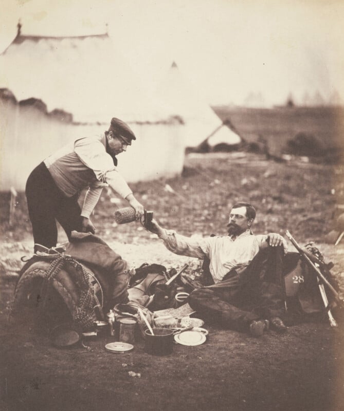 Two men are sitting outside near a rugged campsite. One man, seated on the ground, is handing a bottle to a man standing nearby. They are surrounded by various camping gear, including pots, bags, and a tent in the background.