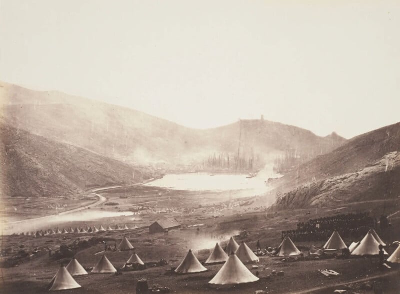 A sepia-toned photo of a military encampment in a valley. Numerous tents are set up on a barren landscape. A lake and hills are visible in the background, with faint outlines of structures or trees. Smoke rises in the distance.