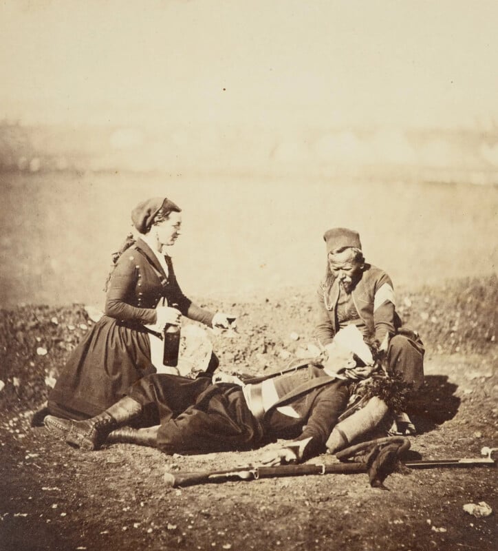 Two women tend to a soldier lying on the ground. One woman holds a bottle and cup, while the other supports the soldier's head. The scene appears to be from the past, with historical clothing and a barren landscape in the background.