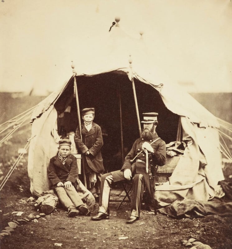 A historical photograph depicts three men in military uniforms sitting outside a canvas tent. Two seated on the ground and one on a chair. The backdrop is a blurred, barren landscape, evoking a wartime setting.