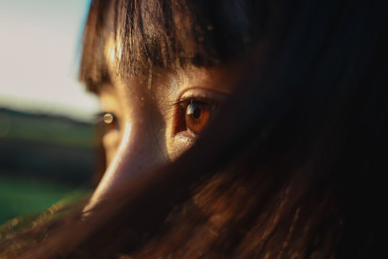 Close-up of a person's eye with long brown hair partially covering their face. The warm lighting highlights the vibrant brown iris and smooth skin, with a blurred natural background.