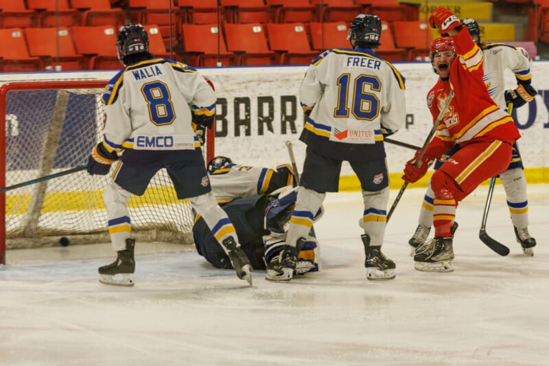 Hockey players in white and blue jerseys defend the goal as a player in a red and yellow jersey celebrates with one arm raised. The goalie is on the ice. The scene takes place on an ice rink with red seats in the background.