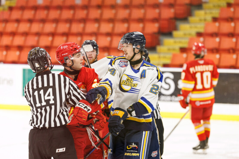 A hockey referee separates two players having an on-ice confrontation. One player in a red uniform and helmet faces another in a white and blue jersey. Additional players and the stands are visible in the background.