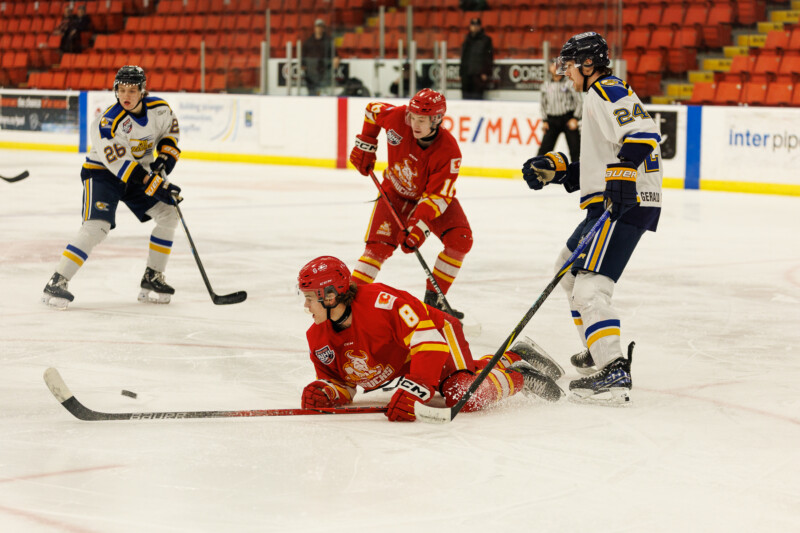 Hockey players in red and blue uniforms are on the ice during a game. A player in red slides on the ice while others look on. The arena has red seating and visible advertisements on the walls.