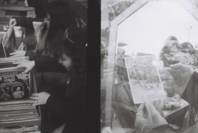 Two women browse through vinyl records in crates on the left side, while on the right, a person holds up a record with a winter scene. The image is in black and white, capturing a vintage, nostalgic atmosphere.