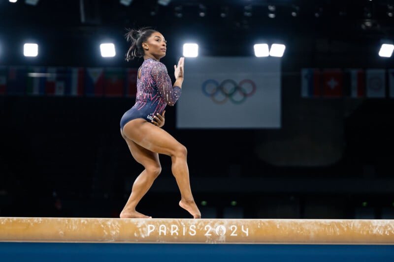 A gymnast in a blue and red leotard performs a routine on a balance beam during the Paris 2024 competition. The Olympic rings and various national flags are visible in the background.