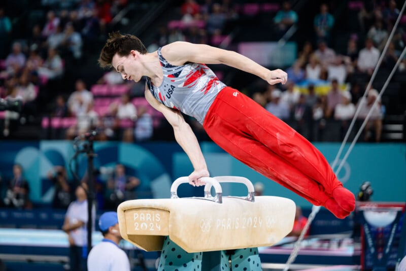 A gymnast wearing a USA uniform is performing a routine on the pommel horse. His body is extended horizontally, showcasing his strength and balance. The background shows an audience and Paris 2024 signage, indicating that this is an Olympic event.