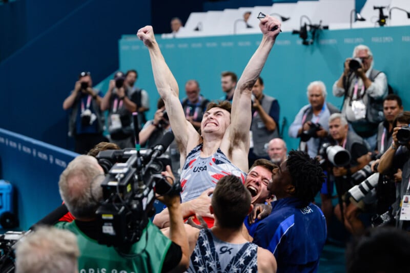 A victorious athlete wearing a USA uniform raises his arms triumphantly as he is lifted by teammates and surrounded by photographers. The scene is one of celebration, with people capturing the moment and sharing in the athlete's joy.