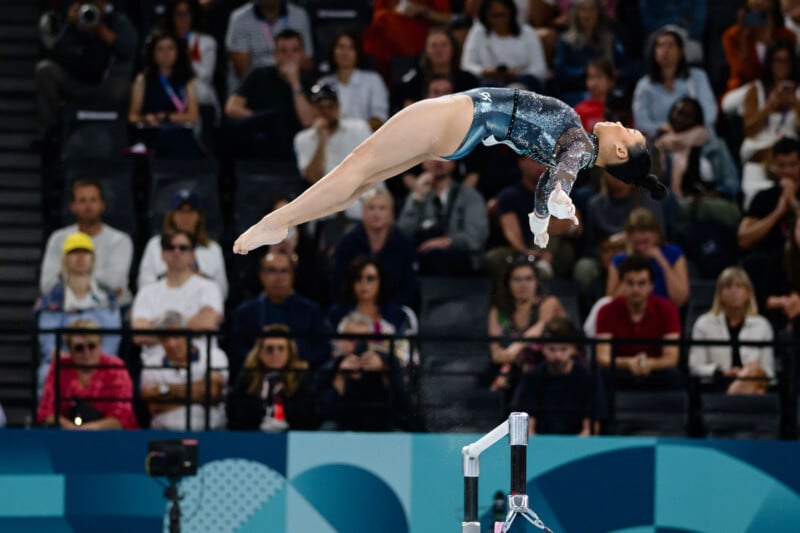 A female gymnast, wearing a sparkling blue leotard, performs a high-flying jump above the uneven bars at a competition. The audience watches intently from the stands in the background.