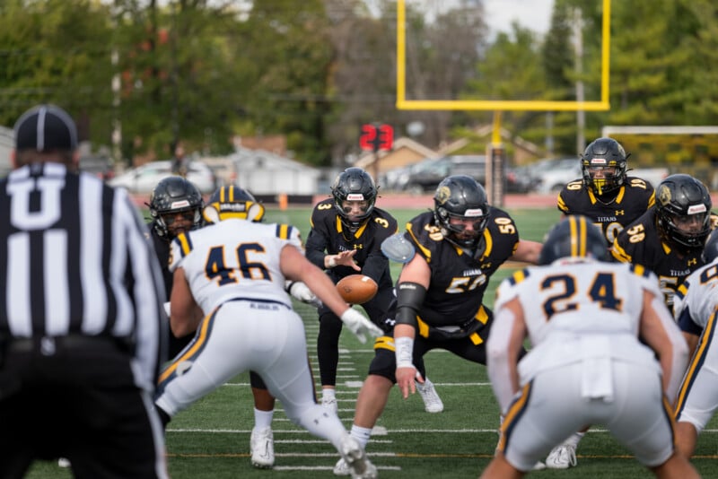 A football game is in action with a player in a black and yellow uniform preparing to throw the ball. The opposing team, in white and yellow, is lined up defensively. The field goal post and a timer are visible in the background.