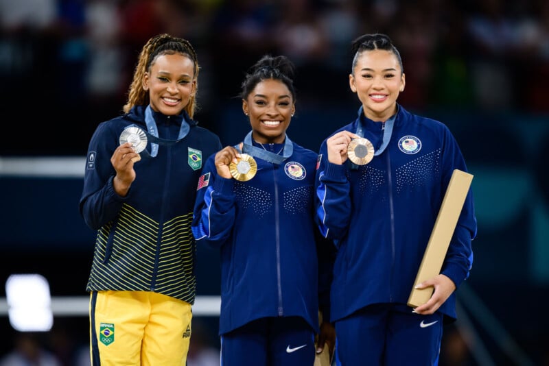 Three female athletes stand on a podium, smiling and holding their medals. The athlete on the left holds a silver medal and wears a yellow and green outfit. The two on the right wear blue tracksuits; one holds a gold medal and the other a bronze.