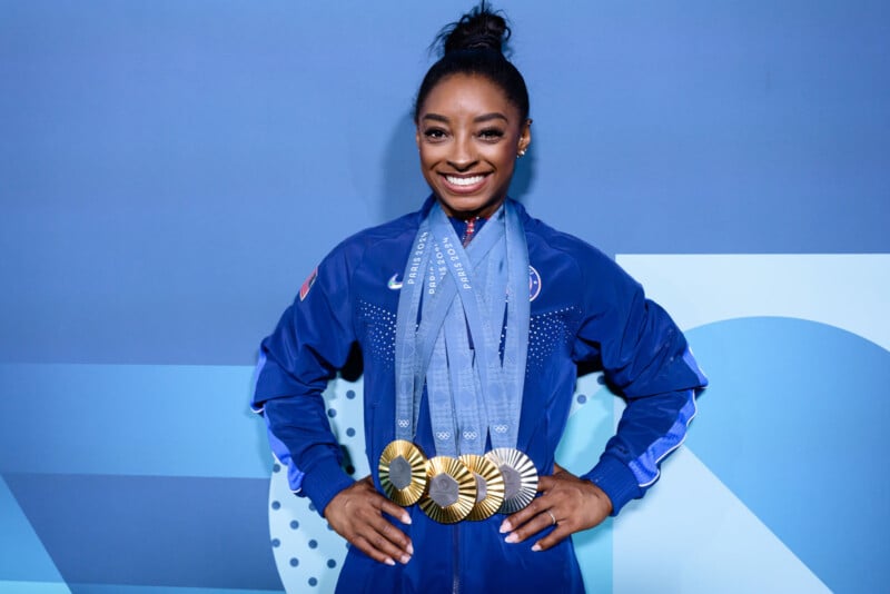 A smiling athlete stands with her hands on her hips, wearing a blue athletic jacket with multiple gold medals hanging around her neck. She has her hair in a high bun and poses against a blue geometric background.