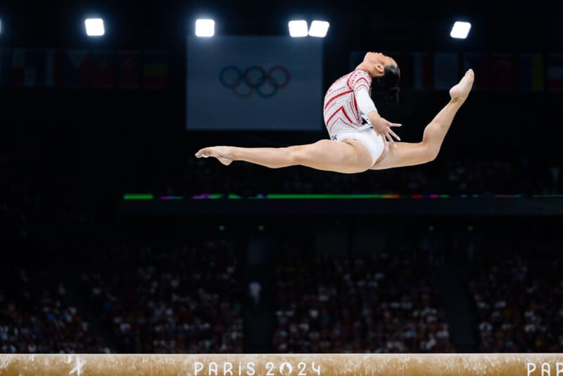 A gymnast in mid-air performing a split leap on the balance beam at an indoor stadium, with a crowd in the background and the Paris 2024 Olympic logo visible.