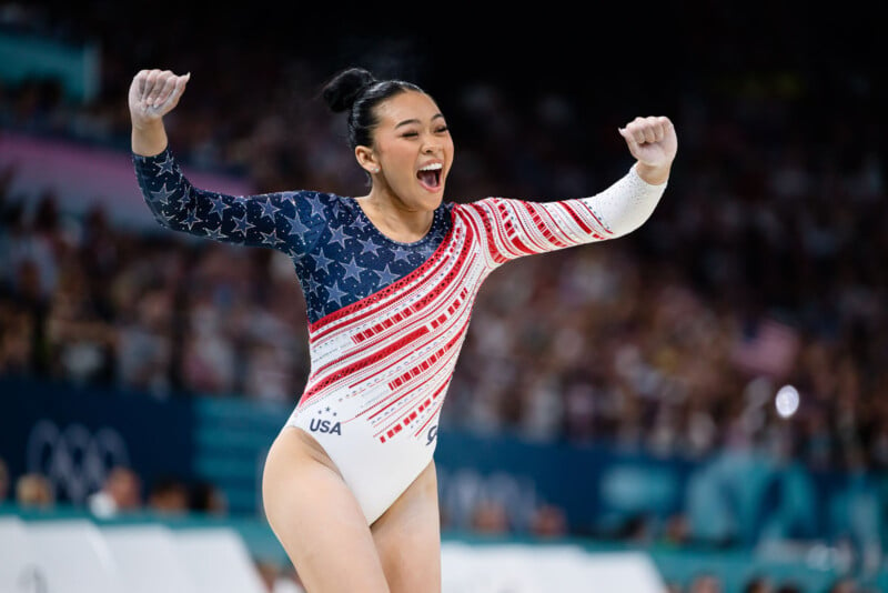 A gymnast wearing a patriotic leotard with USA printed on it celebrates with fists raised. The leotard features blue with white stars on one side and red and white stripes on the other. She is smiling widely with a cheering crowd in the blurred background.