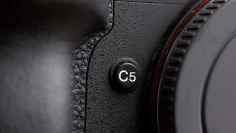 Close-up of a black camera body showing a textured surface and a button labeled "C5" near the grip. The edge of a lens mount is visible on the right.