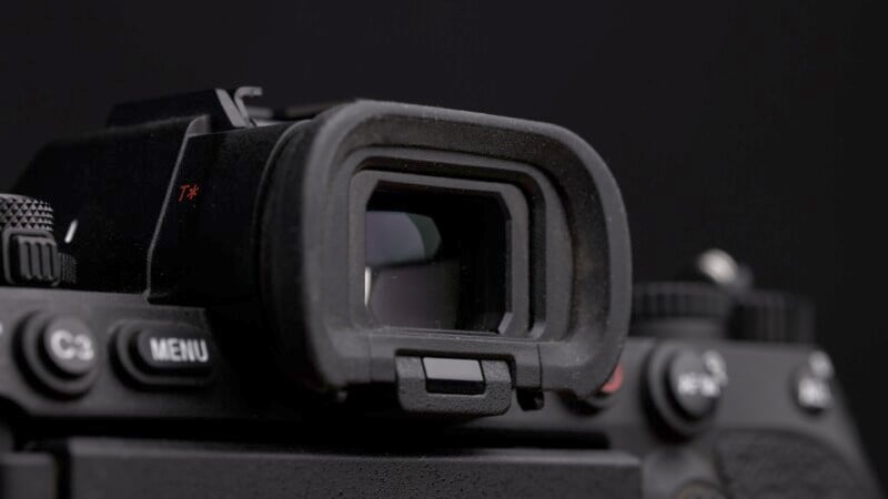 Close-up of a digital camera's viewfinder on a dark background. The viewfinder is surrounded by black rubber, and camera control buttons are partially visible around it.