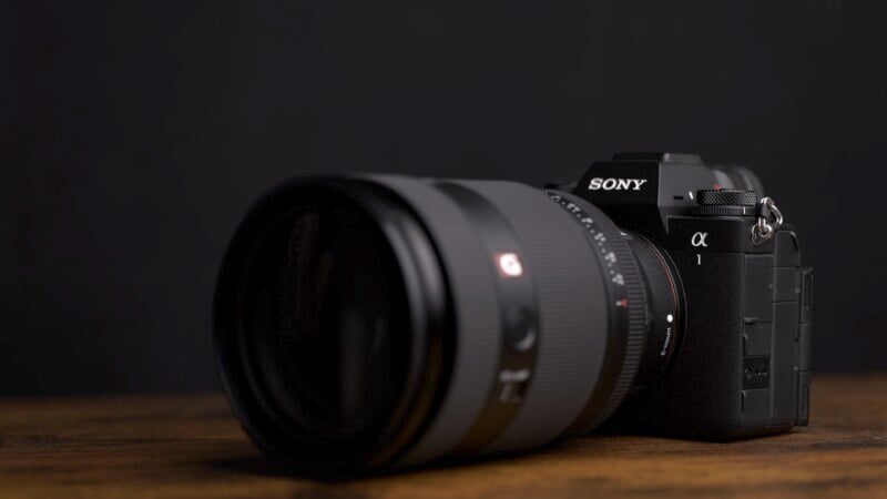 A Sony camera with a large lens is positioned on a wooden surface against a dark background. The camera is angled slightly, showcasing the lens and the brand logo.