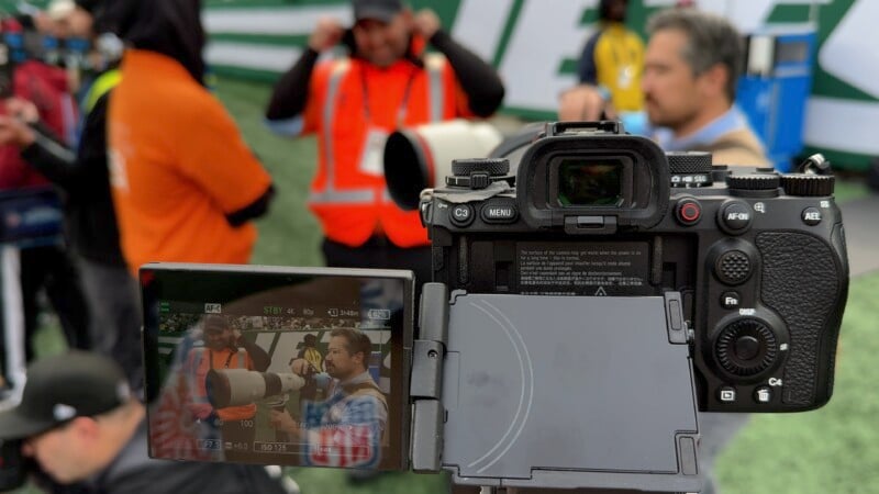 A DSLR camera on a tripod captures an event with people in bright safety vests and jackets. The camera screen displays the image being captured, showing two individuals holding cameras and talking. Blurred stadium seating is visible in the background.