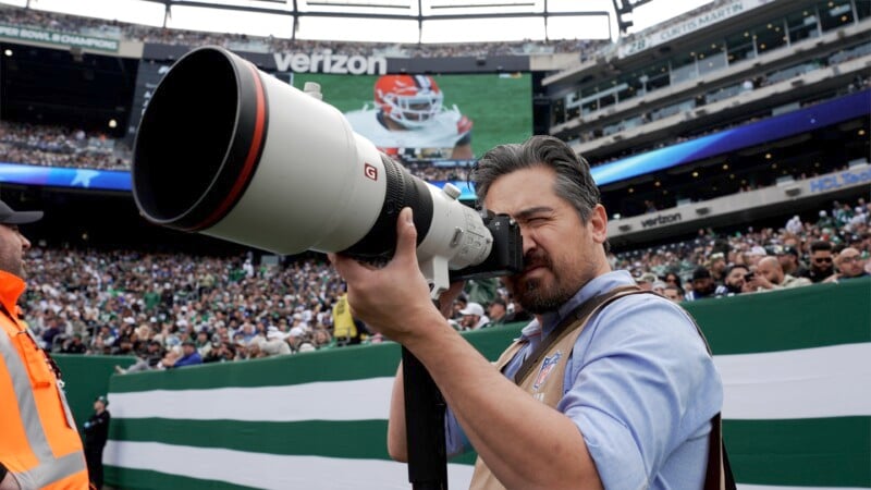 A photographer uses a large telephoto lens on the sidelines of a football stadium. A game is visible on the large screen in the background. The stands are filled with spectators.