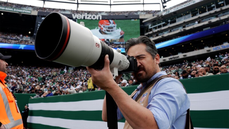 A photographer at a football game uses a large telephoto lens camera. The stadium is crowded with spectators, and a digital screen in the background displays a close-up of a player in action.