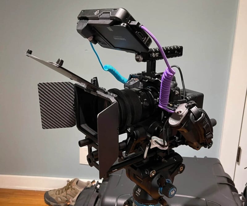 A professional video camera setup is mounted on a tripod, featuring a matte box, external monitor, cables, and other attachments. A pair of shoes is visible on the floor in the background. The scene is indoors with a gray wall and a door to the side.