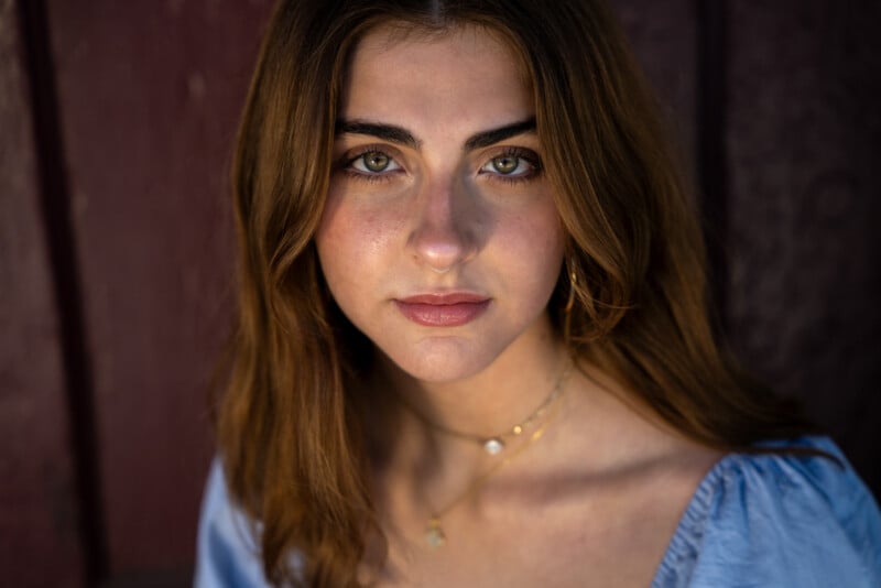 A woman with long brown hair and green eyes looks directly at the camera. She is wearing a light blue top and gold necklaces. The background consists of dark wooden panels.