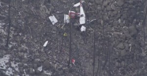 Aerial view of a plane crash site in a rocky, barren area. Debris from the aircraft is scattered among the rocks, with some parts still recognizable. The surrounding area appears charred, indicating possible fire damage.