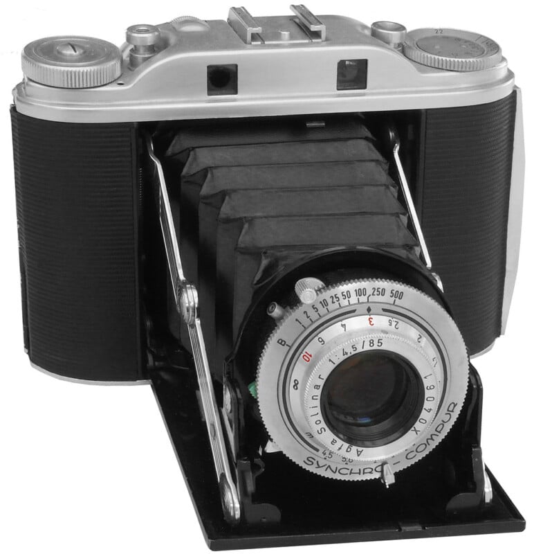 An antique foldable bellows camera with a silver and black exterior. The lens is prominently displayed, featuring markings and numbers. The camera has a vintage, classic design.