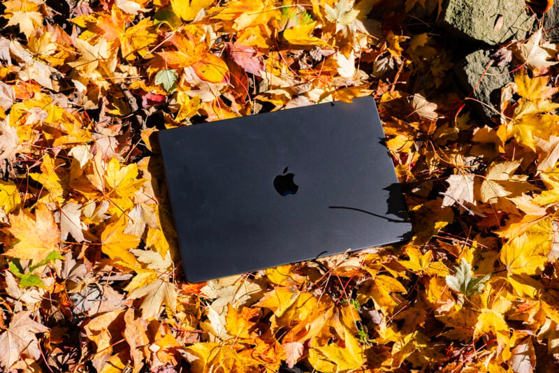 A closed laptop with an apple logo lies on a bed of yellow and orange autumn leaves. Sunlight illuminates the scene, casting shadows on the laptop and leaves.