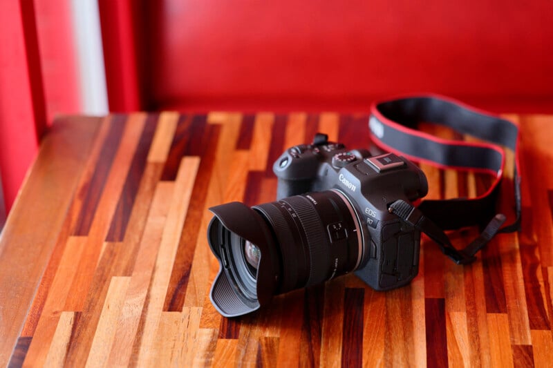 A Canon camera with an attached lens and strap sits on a wooden table with a striped pattern, against a red background.