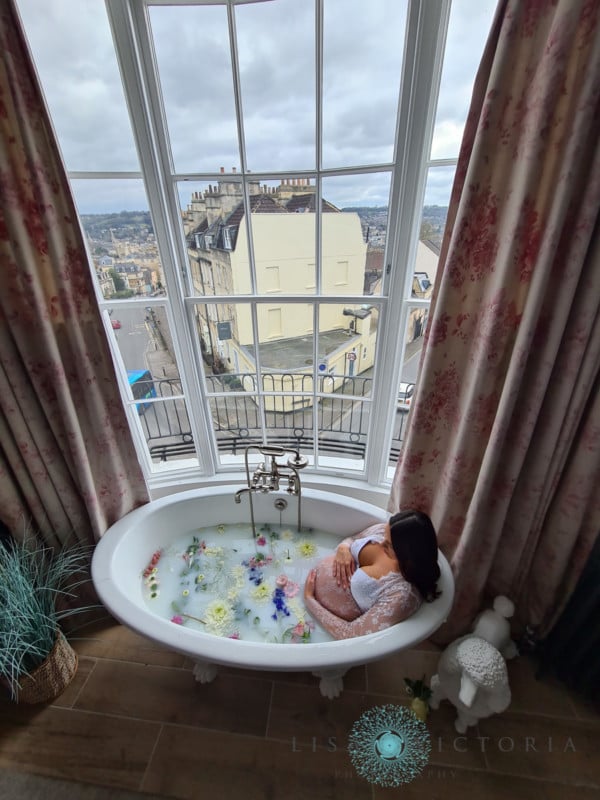 A pregnant mother sits in a milk bath next to a high window