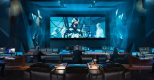 A modern screening room with four people seated at workstations. Multiple monitors display editing software, while a large screen shows a futuristic scene with people in high-tech attire. The room is dimly lit with a blue hue.