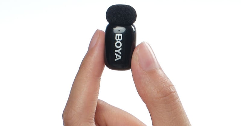 A person holding a small, black BOYA microphone between their thumb and index finger against a plain white background. The microphone has a foam windscreen on top.