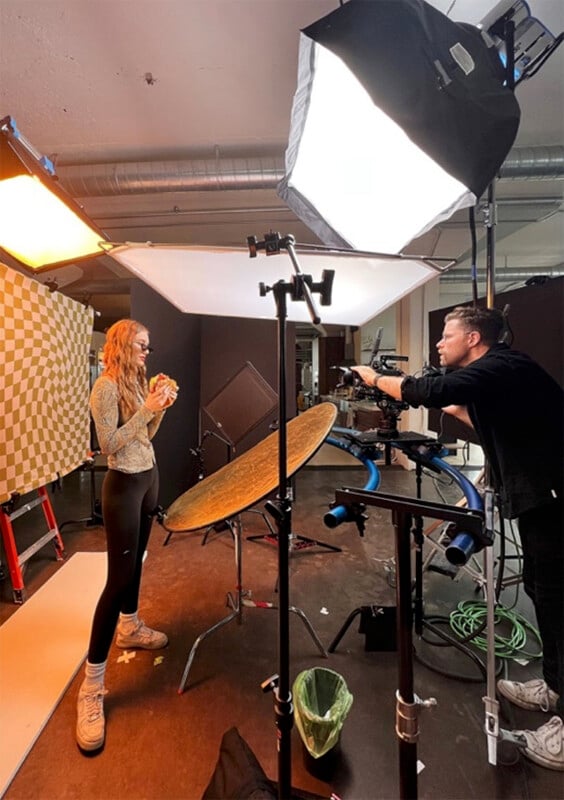 A woman stands in a photography studio, posing with a burger. She is surrounded by professional lighting equipment, including softboxes and reflectors. A photographer is capturing the scene. The setting is industrial and creative.