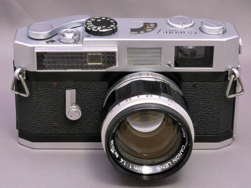 A vintage Canon camera with a silver and black body, textured grip, and a 50mm f/1.4 lens with markings. The top features various dials and buttons. The camera is placed on a plain surface.