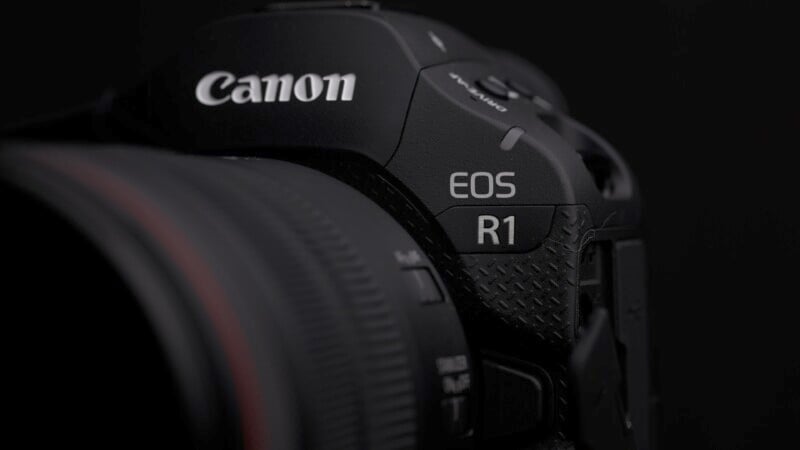 Close-up of a Canon EOS R1 camera, focusing on the camera's branding and body details. The image is predominantly dark, highlighting the camera's sleek design and lettering on the upper right side.