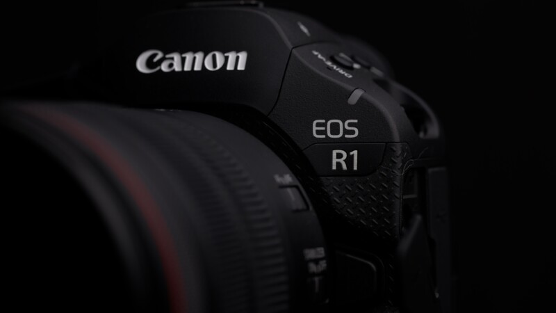 Close-up of a Canon EOS R1 camera. The image focuses on the camera's name and model details, featuring a textured black surface with subtle lighting revealing the design elements.