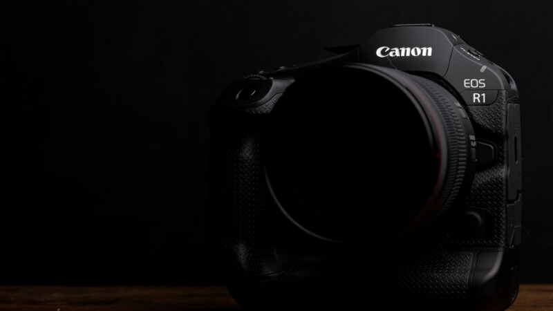 A Canon EOS R1 camera against a dark background, partially illuminated to highlight its textured body and lens. The camera is positioned slightly to the right, with a focus on its logo and model name.